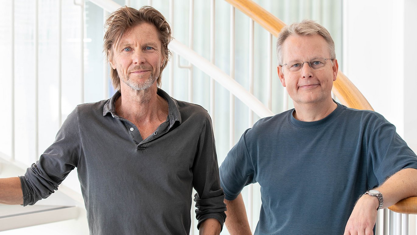 To mænd står indendørs ved en trappe og smiler til kameraet.